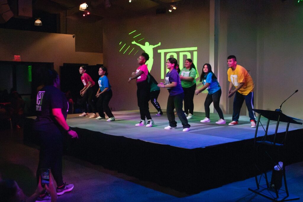 Students in pink, purple, blue, green shirts dancing.