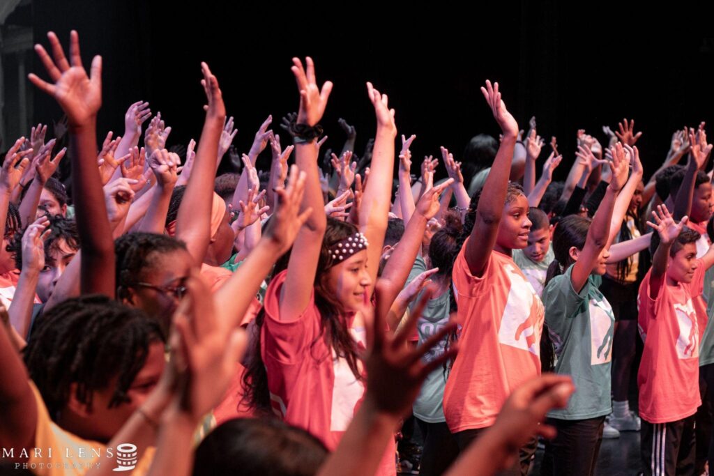 Students in orange, green, red, yellow shirts and black pants dancing on stage.